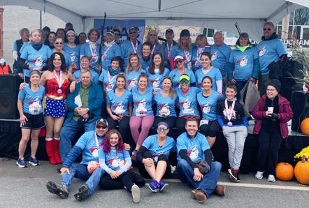Team Winnie's Warriors groups together in their blue team shirts at the 2019 CHaD HERO, sitting on the stage, under a tent. 